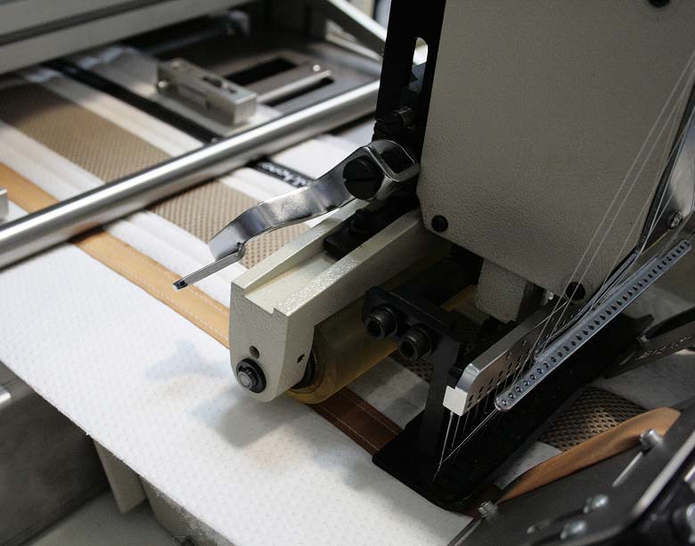 Vista cercana del cabezal de costura multiaguja y el prensatelas, cosiendo y uniendo las diferentes capas de tejido de la platabanda con alta precisión en la máquina industrial.