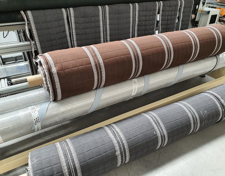 Rollos de tejido de platabanda de colchón acabados, mostrando el acolchado y el corte limpio por ultrasonidos, listos para ser usados en la línea de confección.