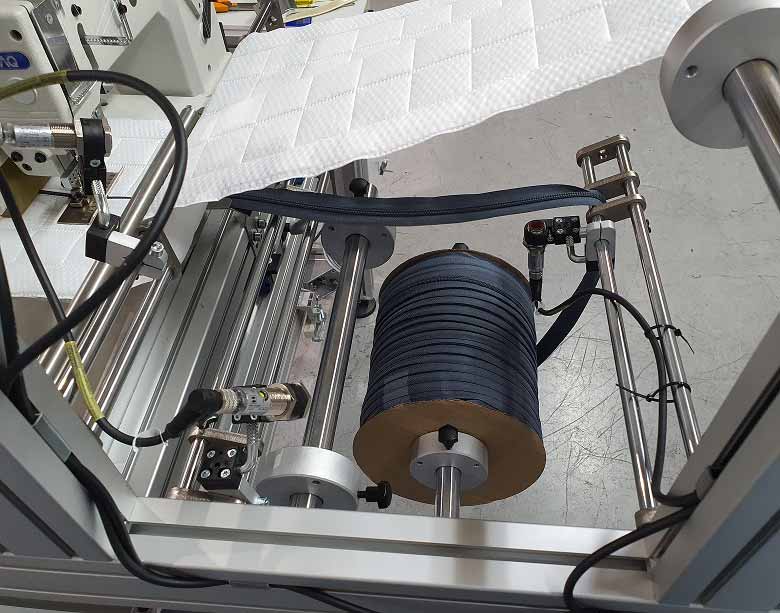 Vista cercana del sistema de alimentación, mostrando el rollo de cremallera central y las dos bandas de tejido de platabanda entrando en la máquina para ser unidas mediante costura.